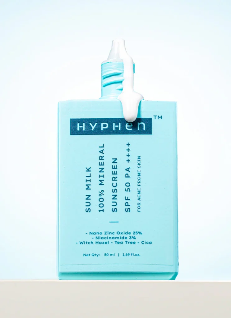 A product shot of Hyphen Sun Milk 100% Mineral Sunscreen SPF 50 PA++++ for acne-prone skin. The square, light blue bottle is shown with a white creamy sunscreen texture dripping from the top. Key ingredients listed on the bottle include 25% Nano Zinc Oxide, 3% Niacinamide, Witch Hazel, Tea Tree, and Cica. Best Mineral Sunscreen